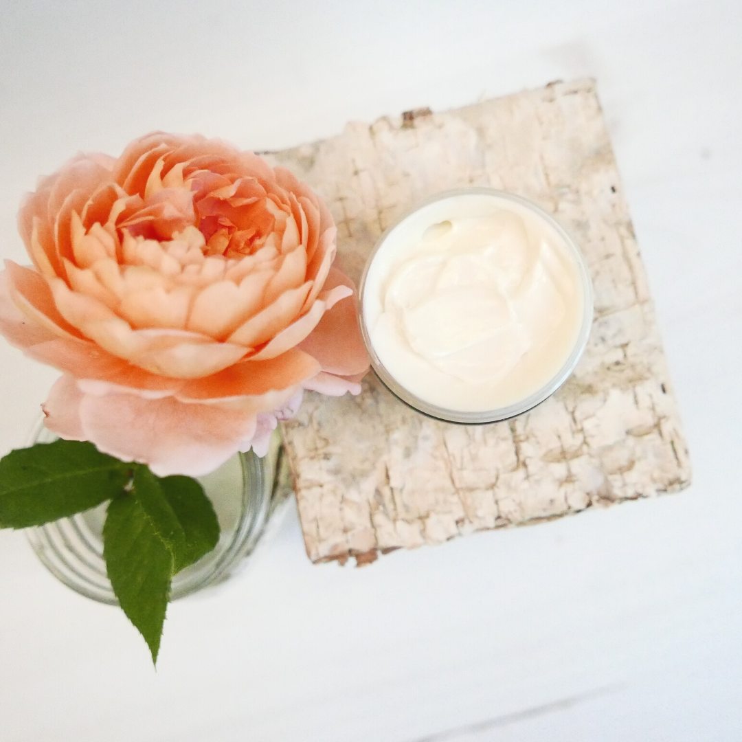 Open calendula facial cream jar showing white cream on a piece of white washed wood next to an orange peony flower.