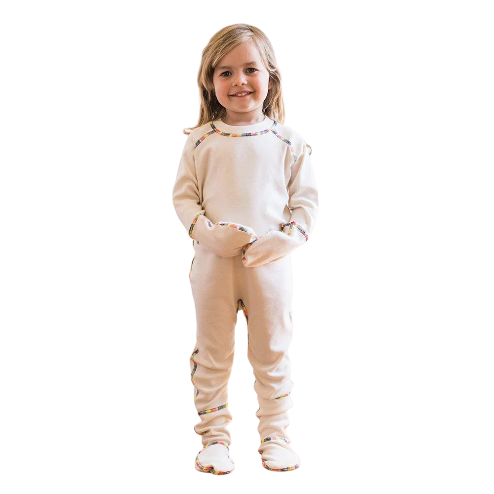 Toddler girl standing and wearing the one piece footie with closed mittens in cream with rainbow piping.