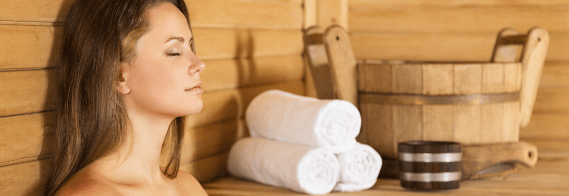 Woman sitting in a wooden sauna with three rolled up white towels and a wooden bucket next to her.