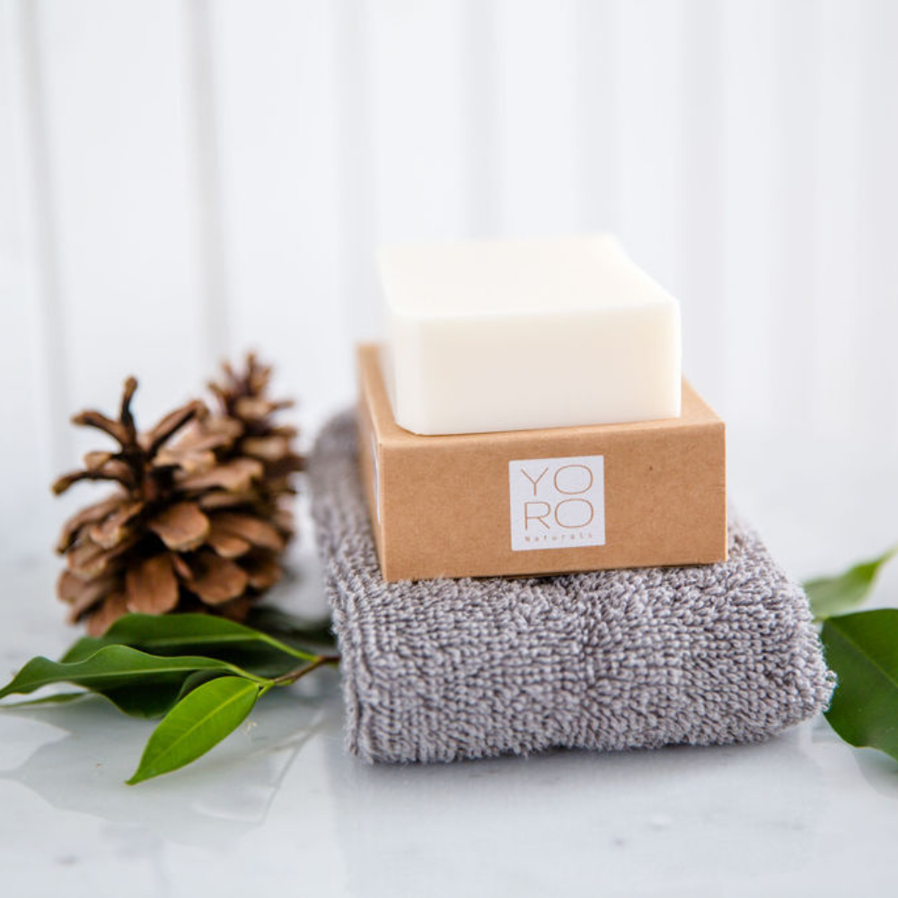 one soap bar on top of it's kraft cardboard box on top of a grey wash cloth and resting on a marble counter next to pinecones and leaves.