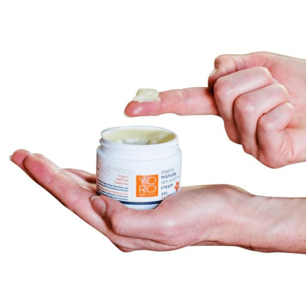 Jar of organic manuka skin soothing cream in palm of hand and a finger dipping into the cream and showing it's texture.